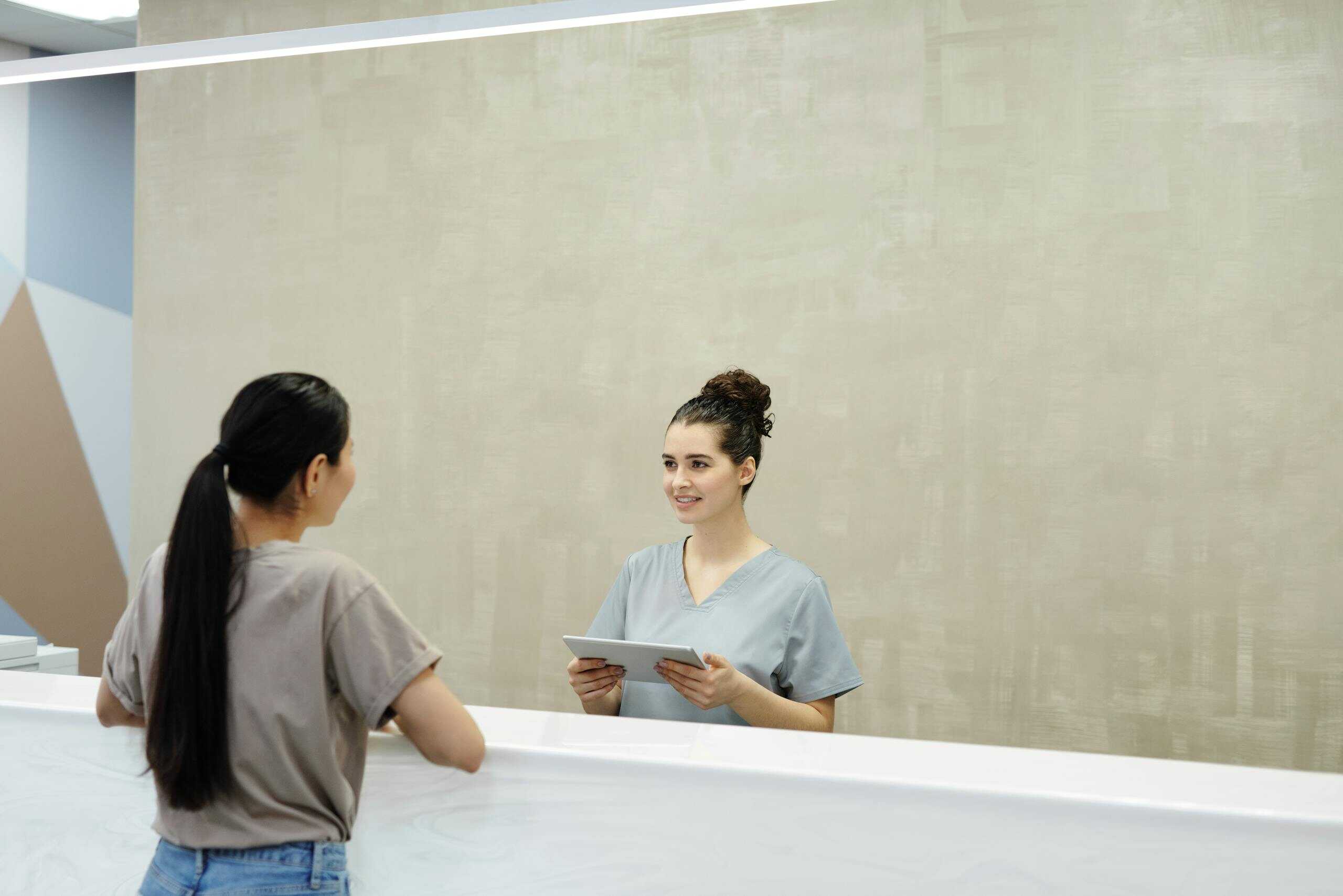 A smiling receptionist with a tablet assists a patient at a medical facility reception desk.